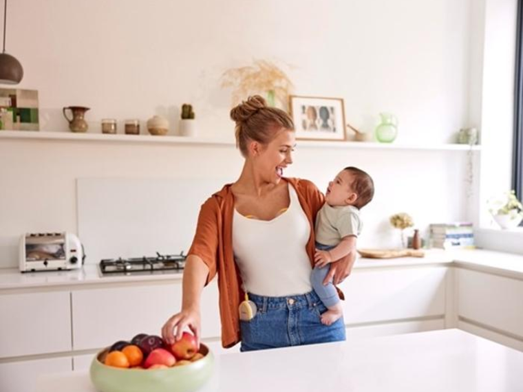Een moeder houdt haar baby vast en eet een snack terwijl ze haar Medela Freestyle™ Hands-free dubbele elektrische borstkolf draagt.