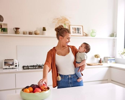 A mom holds her baby and has a snack while wearing her Medela Freestyle™ Hands-free double electric breast pump.