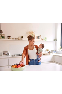 A mom holds her baby and has a snack while wearing her Medela Freestyle™ Hands-free double electric breast pump.