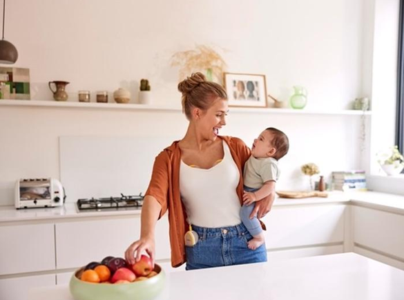 Een moeder houdt haar baby vast en eet een snack terwijl ze haar Medela Freestyle™ Hands-free dubbele elektrische borstkolf draagt.