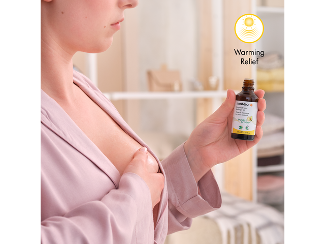 A mom holds a bottle of Medela's Organic Massage Oil in her hand after applying it to her breast. Image text reads: "Warming relief"