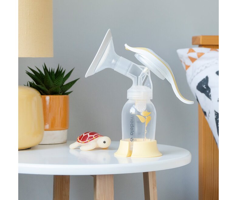 Een Medela Harmony™ handmatige borstkolf op een nachtkastje met een lamp, een plant en speelgoed.