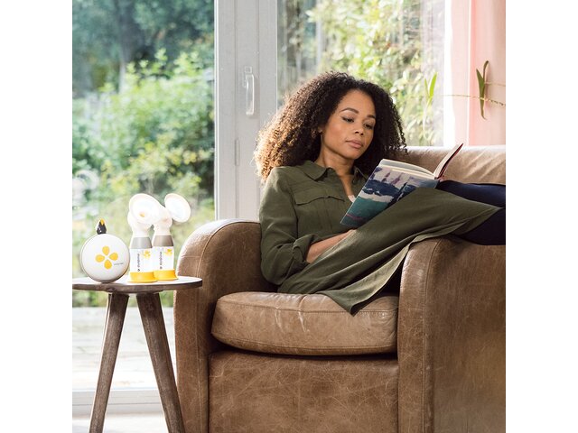 A woman relaxing and reading at home with her Medela Swing Maxi™ Hands-free Electric Breast Pump and classic collection containers next to her.