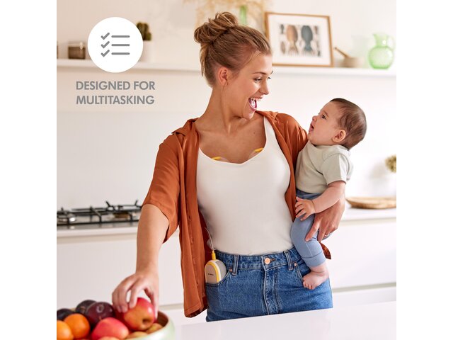 A mother holds her baby and has a snack while wearing her Medela Freestyle™ Hands-free double electric breast pump. Text: “Designed for multitasking”