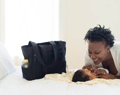 Baby laying on bed next to a Medela pumping bag.  Woman is smiling down on baby.