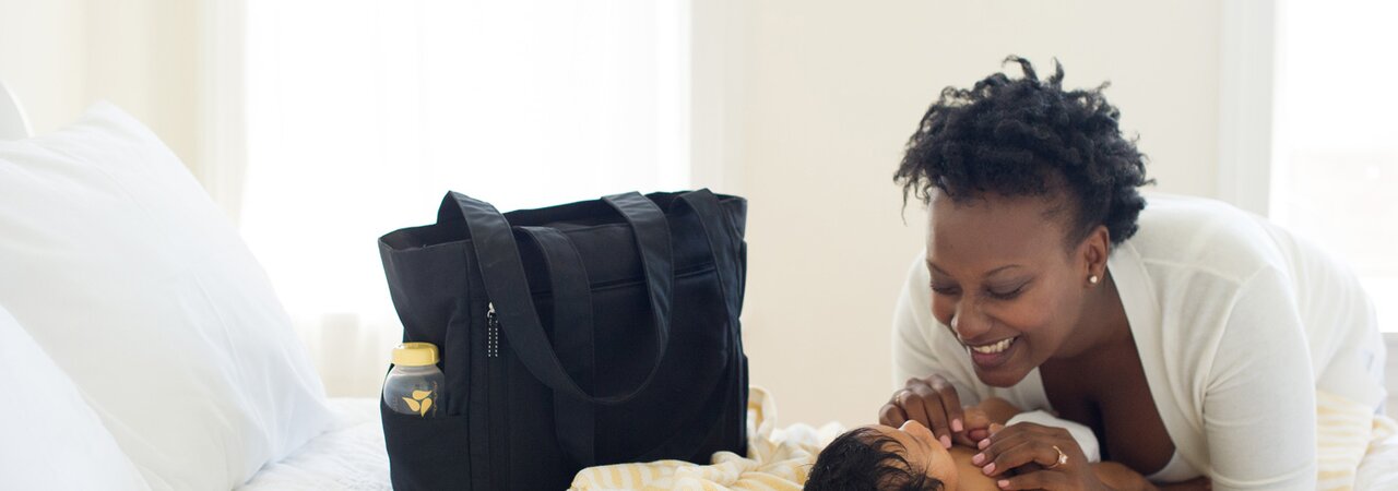 Baby laying on bed next to a Medela pumping bag.  Woman is smiling down on baby.