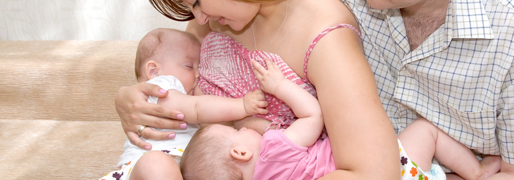 Een moeder voedt haar tweeling in tandem terwijl de vader naast hen zit en een van de baby's ondersteunt.