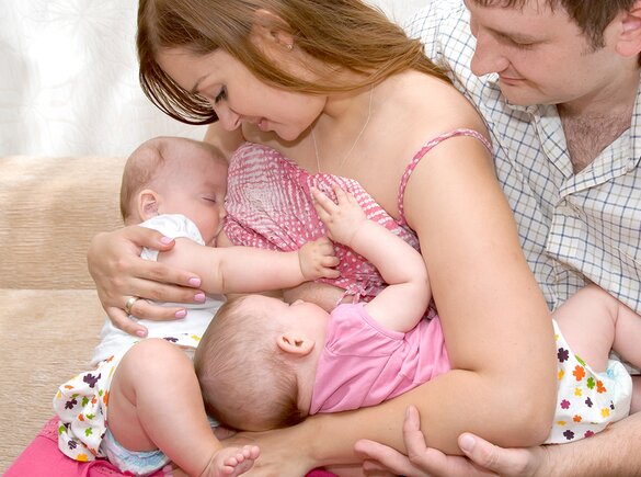 Een moeder voedt haar tweeling in tandem terwijl de vader naast hen zit en een van de baby's ondersteunt.