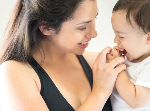 Een baby probeert zijn handjes in zijn mond te stoppen en zijn moeder houdt speels zijn handjes vast, waardoor ze glimlachen.
