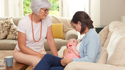 A mother breastfeeds her baby at home in the living room while the baby's grandmother supports her.