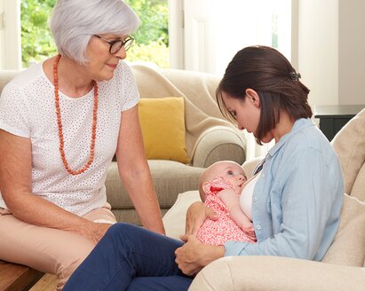 Una madre amamanta a su bebé en el salón de su casa mientras la abuela del bebé la apoya.