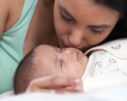 Ein Baby liegt auf einem Bett und die Mutter beugt sich über das Baby und küsst es auf die Wange