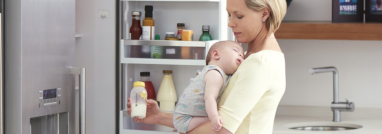 Uma mãe segura o seu bebé na cozinha enquanto prepara leite extraído refrigerado para uma refeição.