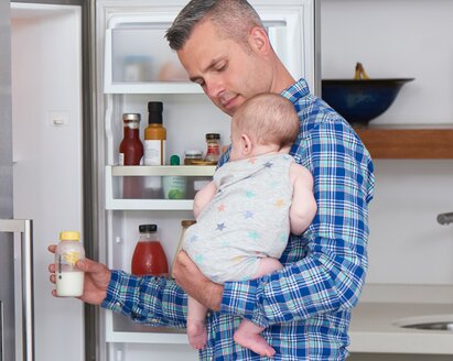 Um pai segura o seu bebé na cozinha enquanto tira um biberão do frigorífico para o aquecer para a alimentação.