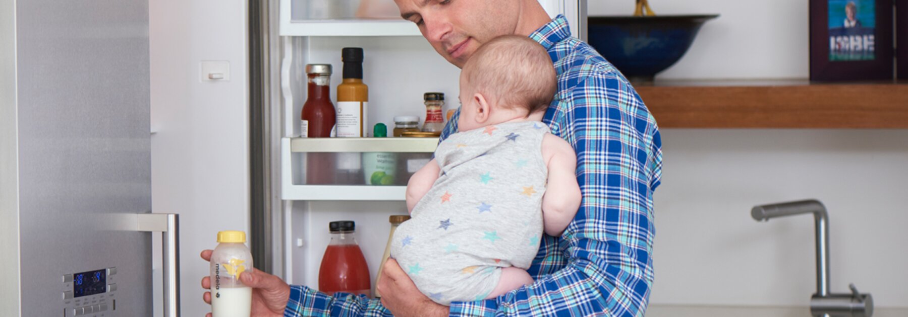 Ein Vater hält sein Baby in der Küche, während er eine Flasche aus dem Kühlschrank nimmt, um sie für die Fütterung aufzuwärmen.