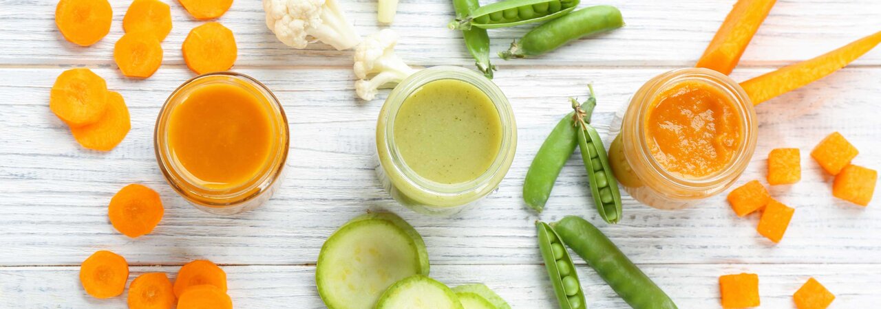 Ein Tisch mit drei gefüllten Gläsern mit Babynahrung, umgeben von geschnittenem Gemüse: Karotten, Erbsen, Blumenkohl, Gurken und Kürbis.